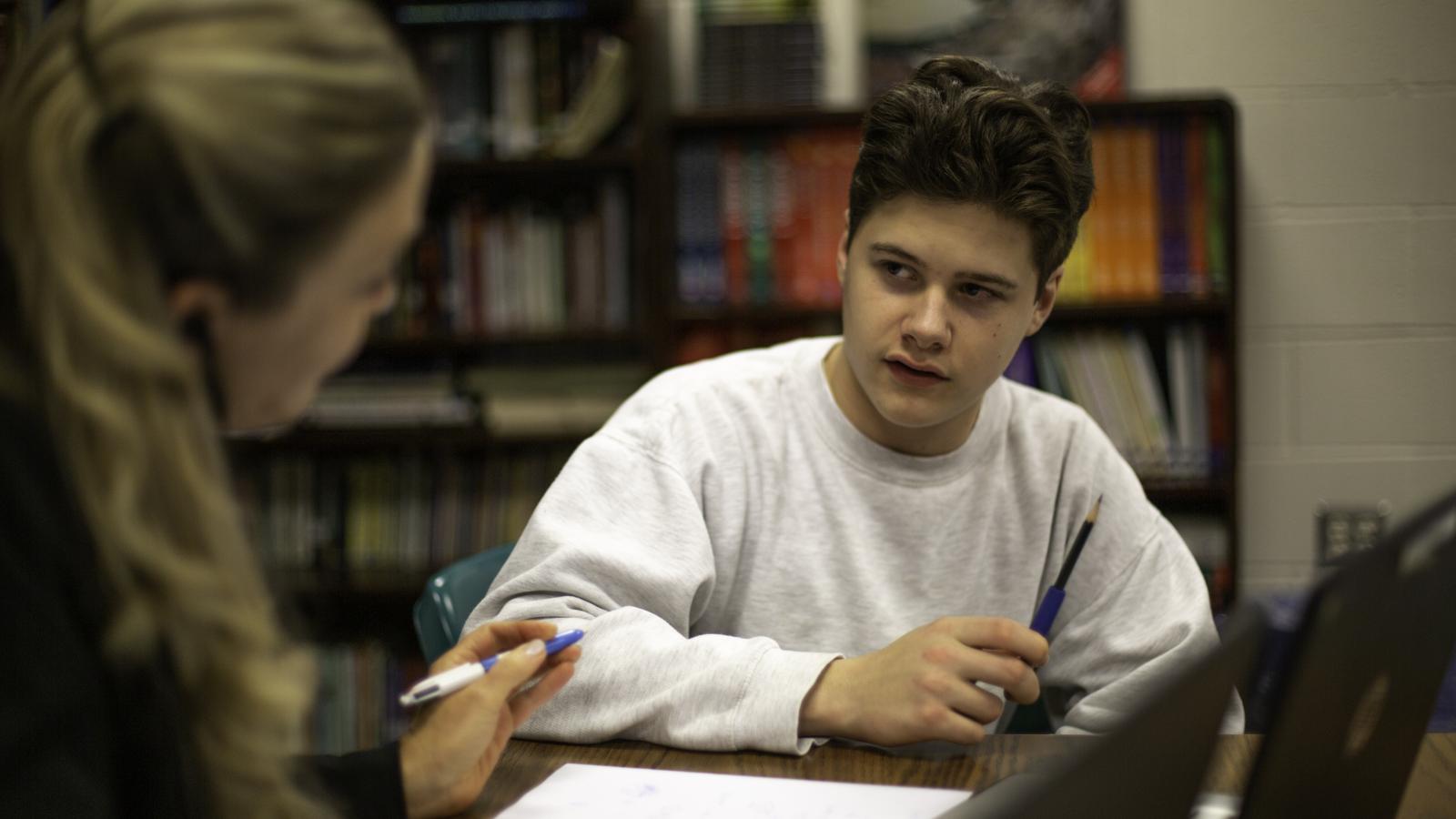 Teenage student seated at a desk, speaking with a teacher