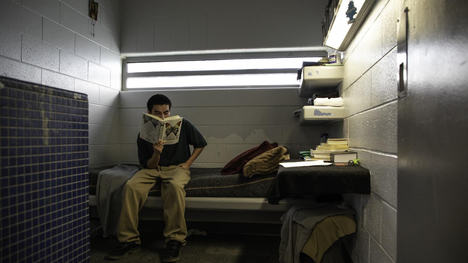 Teenager sitting on a bed in a juvenile facility cell, reading a book